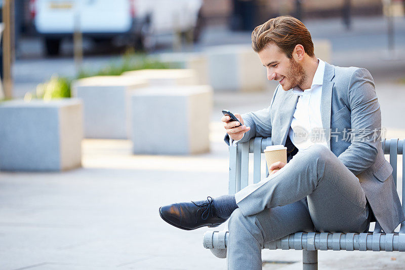 商人在公园长椅上用手机喝咖啡