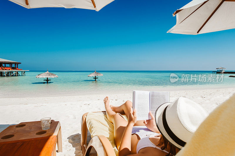 一名女子在热带岛屿的阳伞下躺在日光浴床上看书