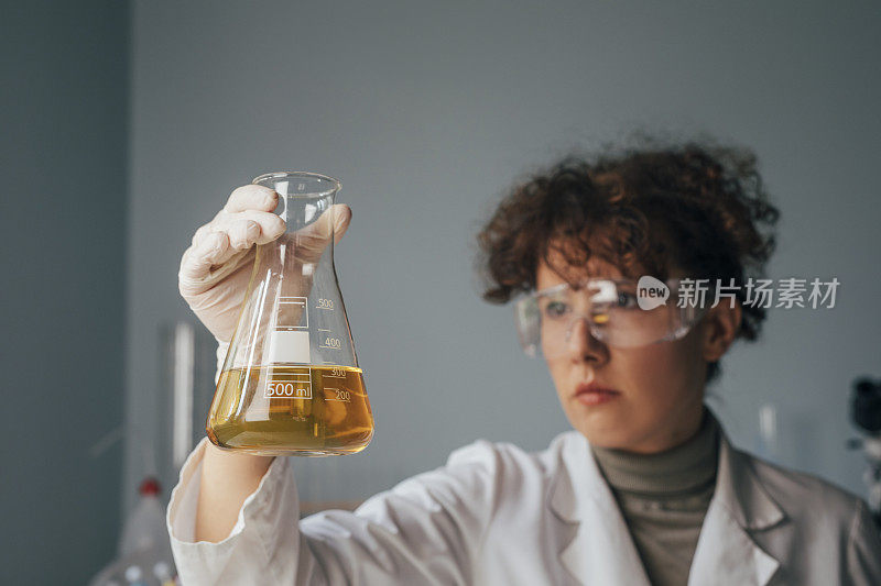 穿着防护装备的年轻女科学家观察锥形瓶中的黄色液体