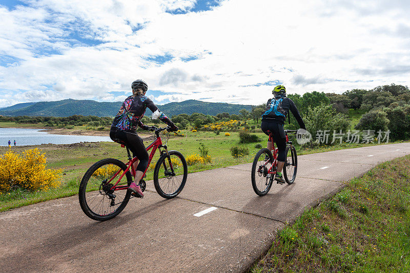 两个年轻人骑着自行车在乡间小路上做运动，以夏日大自然为背景