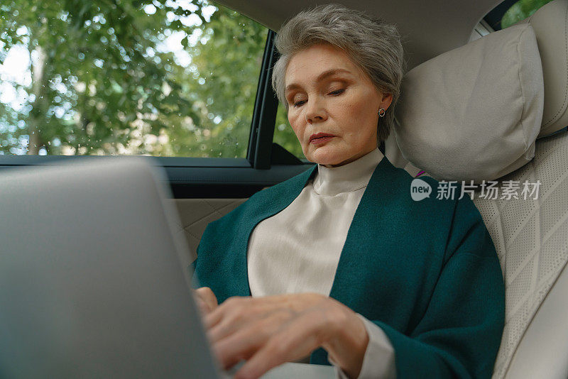一位漂亮的高级商务女性，时尚的发型和专业的妆容，看着她膝上的笔记本电脑坐在一辆昂贵的汽车的乘客座位上