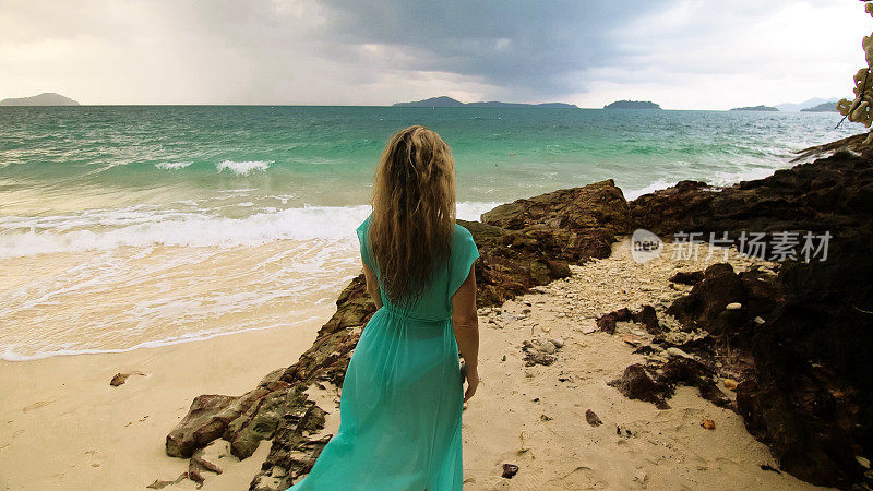 一名女子走到沙滩上暴风雨多云的海洋。女孩在蓝色泳衣连衣裙束腰外衣。概念海上休憩，热带海岸线旅游度假