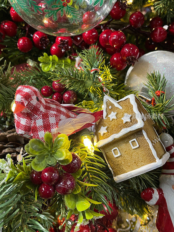 人造的图像，姜饼屋装饰着白色的糖霜和红色和白色的条纹鸟装饰与松果和红色浆果在人造圣诞树的树枝，云杉针，模糊的白色精灵灯的背景