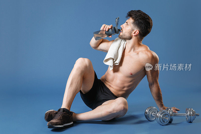 全身像年轻的健身运动强壮的家伙裸露的胸部运动员孤立在蓝色的背景。锻炼，运动，动机，生活方式的概念。坐在地板上，用毛巾搭在肩膀上喝水。