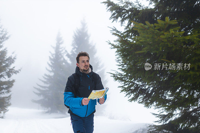 年轻的男性徒步旅行者穿着蓝色的夹克，在雾蒙蒙的雪山上走着，手里拿着地图