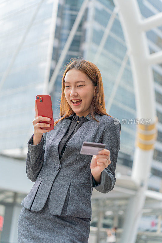 一名年轻的亚洲女商人穿着灰色的办公套装，在商业区用她的智能手机使用信用卡进行在线交易，她微笑着自信