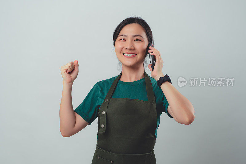 一名穿着围裙的亚洲女子开心地微笑着，用智能手机打电话给客户确认订单。