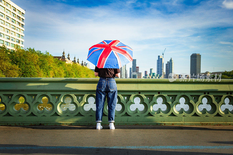 在伦敦市中心的威斯敏斯特桥上，一名女子手持英国国旗伞眺望风景