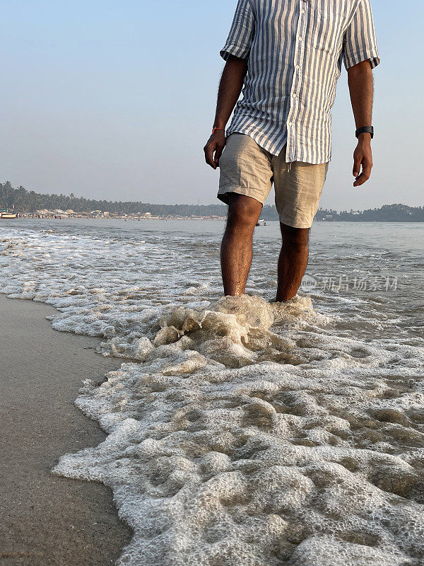 印度果阿帕洛莱姆，黄昏时分，一名无法辨认的男子穿着短裤和条纹短袖衬衫站在沙滩上，度假游客赤脚走在海边，这是前景的焦点