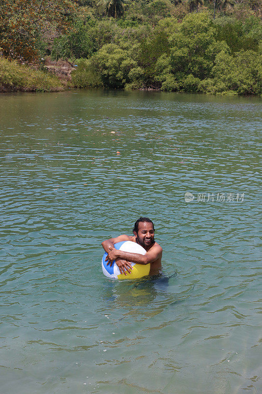穿着冲浪短裤的印度男子站在齐腰深的泻湖平静的水中，抱着漂浮的彩色塑料沙滩球，热带岛屿天堂，林地和棕榈树的景色，前景的焦点