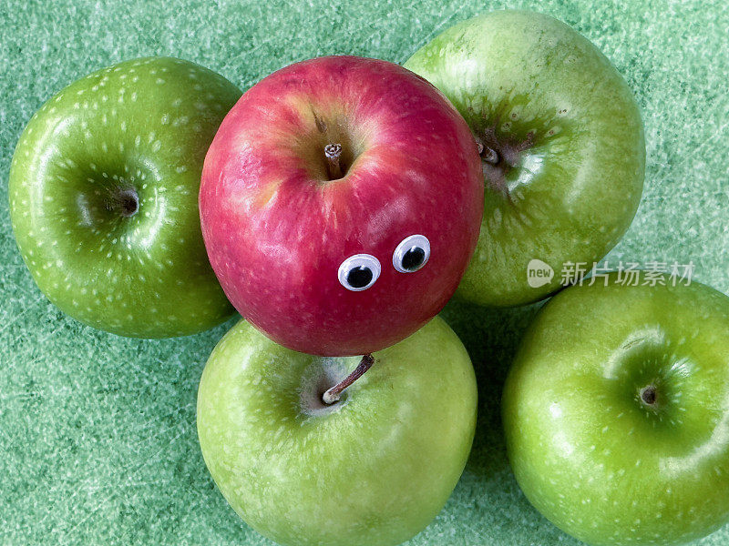 在一组绿色的史密斯苹果上，用红苹果做成的卡通人物的形象，有光泽的，有斑点的水果皮，斑驳的绿色背景，抬高的视角，儿童健康饮食，幽默的概念