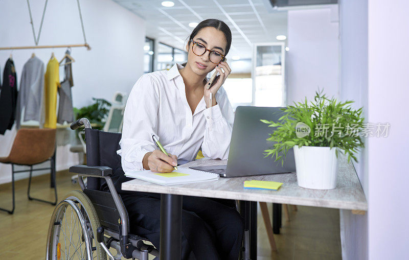 一个漂亮的女孩坐在轮椅上，用她的笔记本电脑工作，用她的智能手机在一个共同工作中心打电话