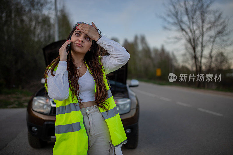 年轻的压力很大的女子站在她抛锚的汽车旁边，用手机求救