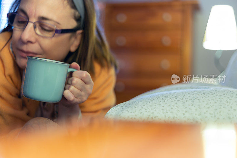 成熟的女人戴着眼镜躺在床上，端着一杯茶、咖啡或输液。