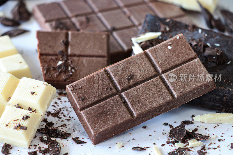 全帧图像的巧克力棒，牛奶巧克力块，白巧克力方块和黑巧克力块与磨碎的刨花在白色大理石效果的背景，重点在前景