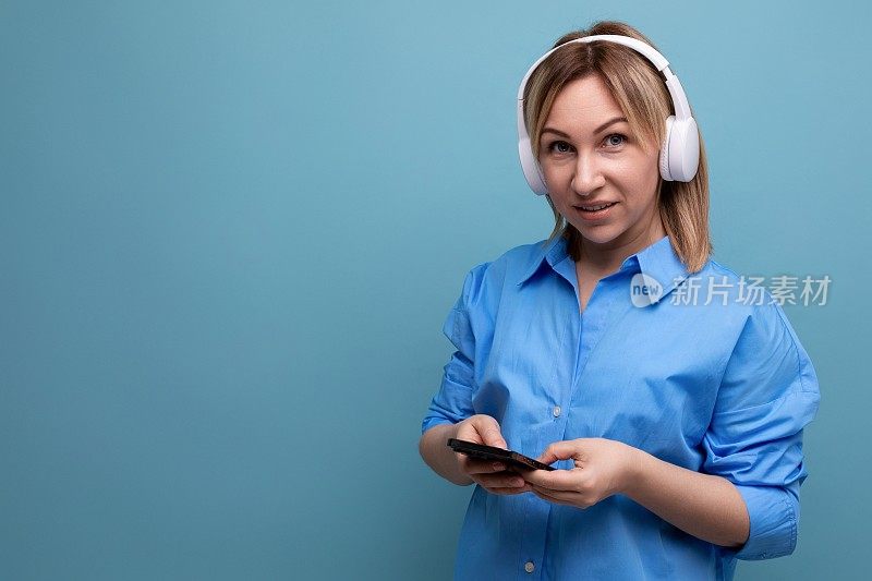 宽屏照片，一个迷人的欧洲女孩穿着休闲衬衫，戴着白色的大耳机，在蓝色的孤立背景上听音乐