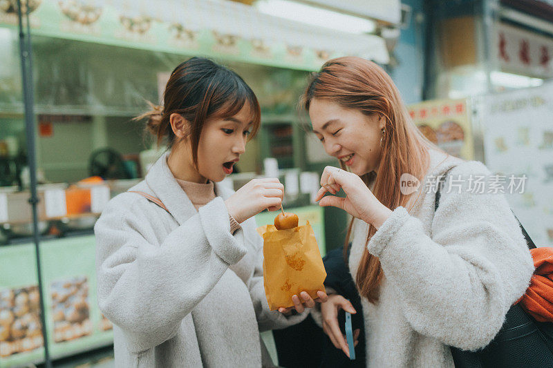 两个亚洲年轻女子正在一起享用一种休闲小吃——鱼丸。他们正站在一个食品摊贩的前面