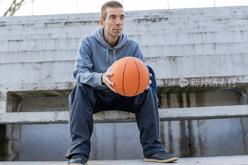 一名男子拿着篮球坐在篮球场旁边的台阶上，赛后休息。