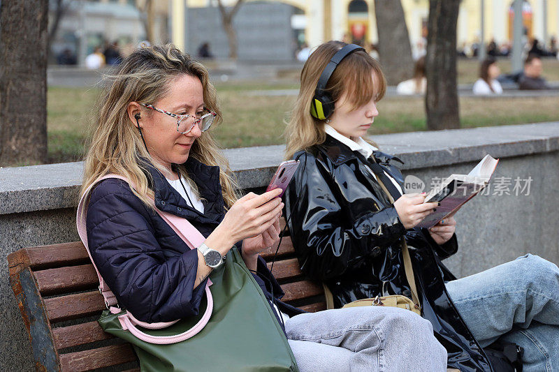 人们坐在街边的长椅上拿着智能手机和纸质书