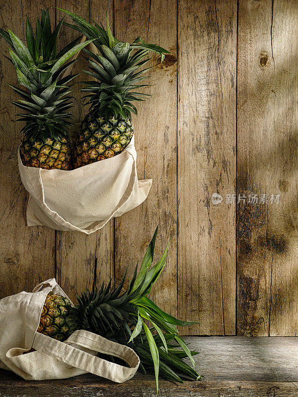 新鲜的热带菠萝装在可重复使用的棉袋里，放在一张质朴的木桌上，背景是饱经风霜的旧木墙。