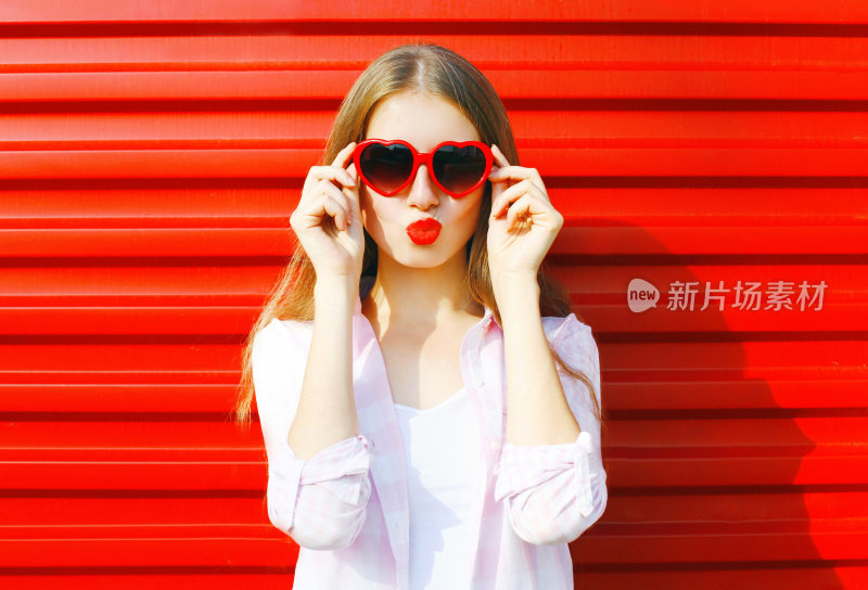 肖像漂亮的年轻女子在红色心形太阳镜在彩色的背景吹嘴唇