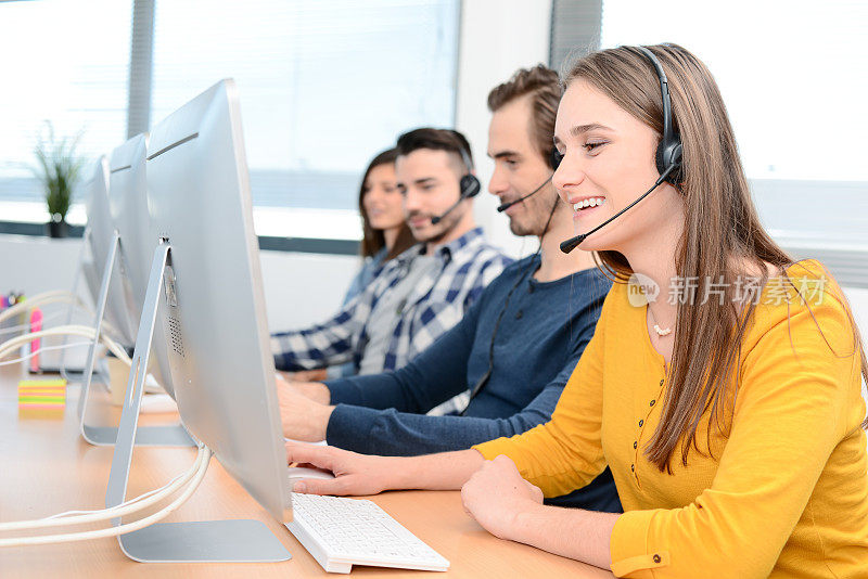 一个美丽开朗的年轻女性电话接线员戴着耳机在桌面电脑上工作，在客户服务呼叫支持热线业务中心，团队工作人员在后台