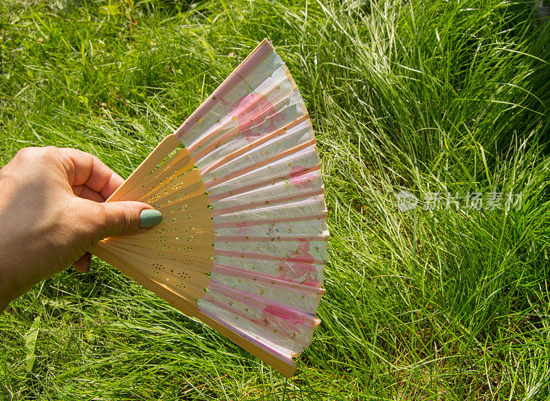 女用手拿着一把打开的竹扇，放在草地的背景上，复制空间