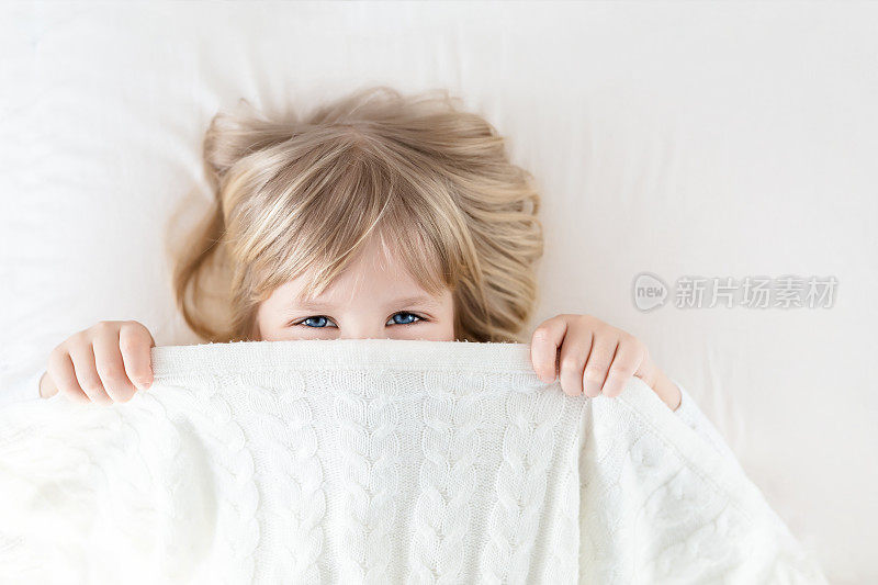 小女孩从床上的毯子里往外偷看。可爱的孩子微笑着，藏在针织被子里。淘气的眼神。捉迷藏。孩子们玩得很开心。快乐的童年概念