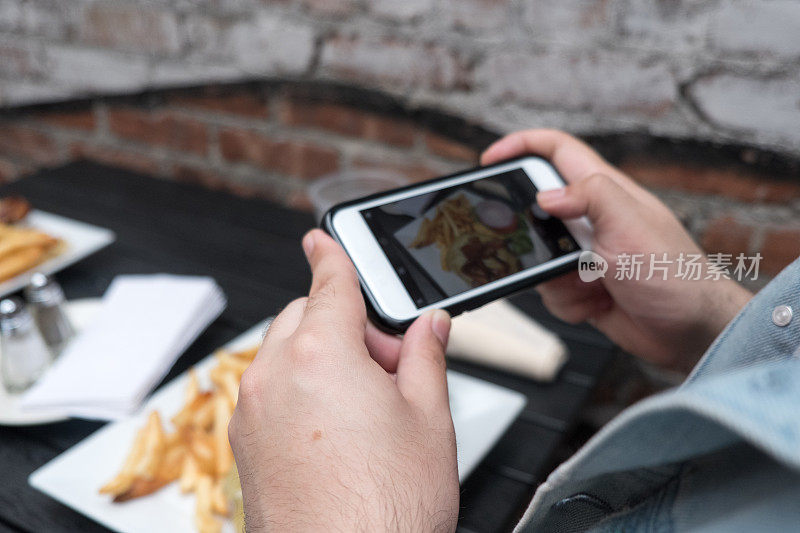 一名男子拿着手机给他的食物拍照。智能手机食品摄影。在一家户外酒吧用智能手机拍下汉堡和炸薯条的照片。