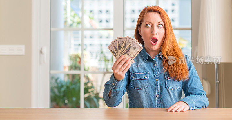 红发女子手里拿着一美元钞票在家里吓得一脸吃惊，又怕又激动又害怕的表情