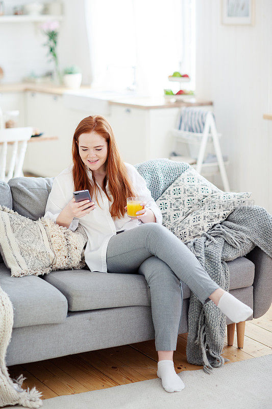 一个红头发、面带微笑的年轻女子坐在家里的沙发上，用智能手机看短信。她手里拿着一杯橙汁
