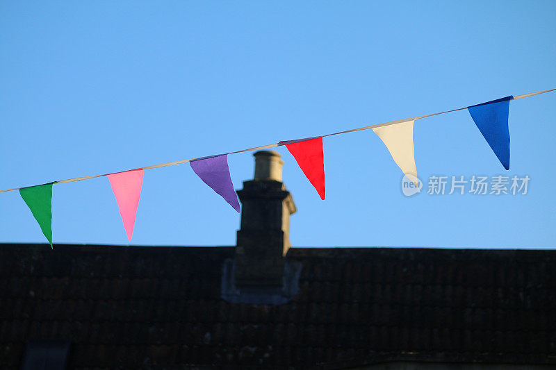 彩虹彩旗悬挂于街道上的彩色彩旗图像，在蓝天、烟囱、烟囱和屋顶上悬挂彩旗，为一年一度的街头派对嘉年华庆祝活动拍摄彩旗照片