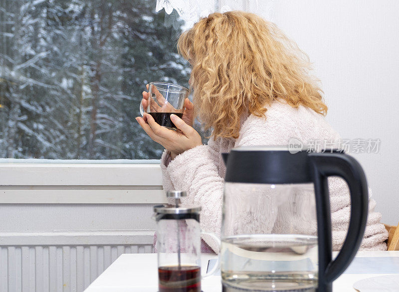 一个年轻的白人女子一边喝着咖啡，一边望着窗外。早餐在厨房。