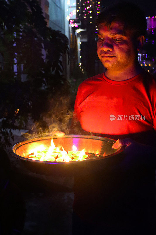 一名印度男子拿着一个装有排灯节油灯的塔利(金属托盘)，装满油的黏土油灯，在黑暗中闪烁的火焰，排灯节的灯庆祝，高架的视野，聚焦前景