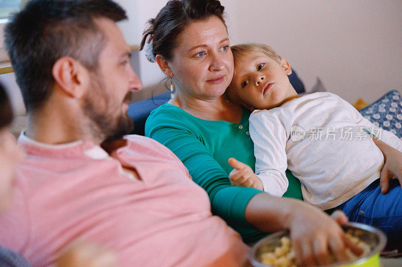 母亲带着蹒跚学步的孩子在她的腿上吃零食，和她的家人一起看电视