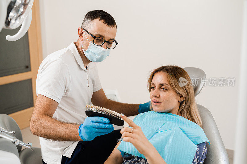 牙科医生在牙科诊所为女性病人示范牙齿美白的颜色深浅。牙医。妇女看着贴面或种植牙配色样本在医生手中。