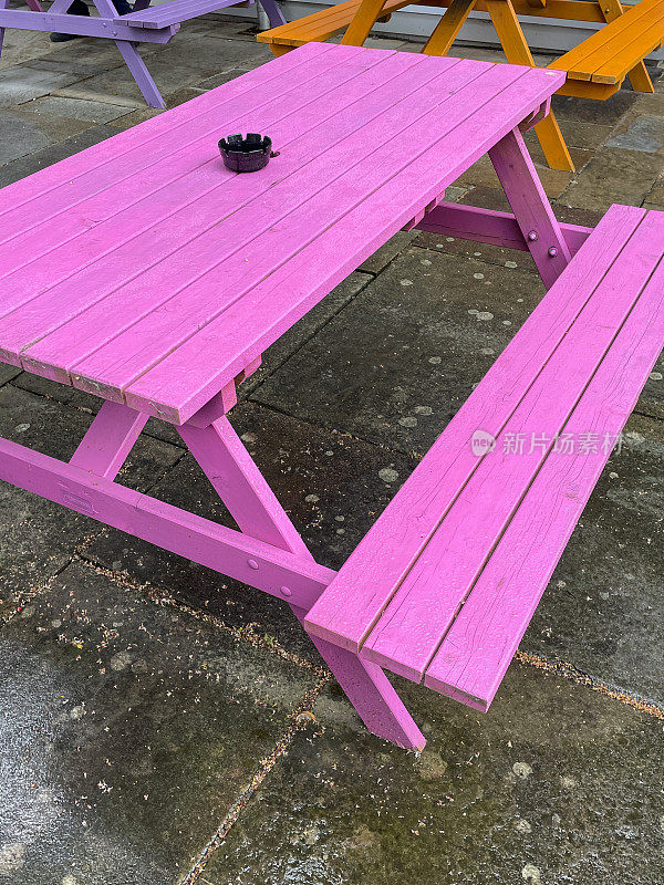 彩色绘画的特写图像，木制户外野餐桌子和长椅铺在露台区域，粉红色，橙色和紫色的户外家具，重点前景