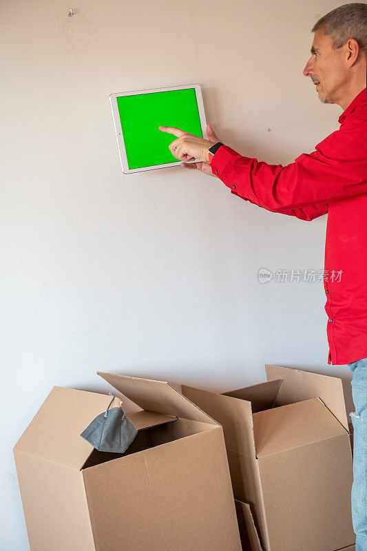 搬到地板上满是纸板箱的新家。智能家居和科技概念，男性的手指向平板电脑与绿色屏幕集成在房子的室内墙壁。