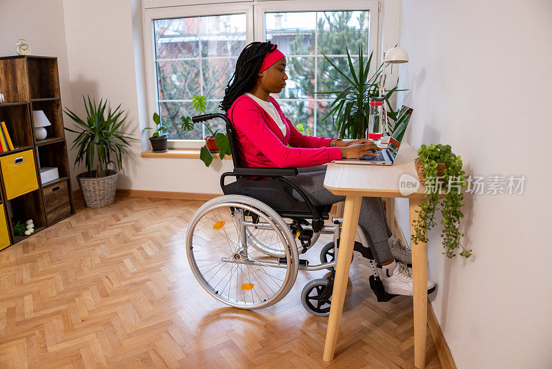 一个黑人妇女坐在轮椅上用笔记本电脑工作的侧面视图，从她的家庭办公室