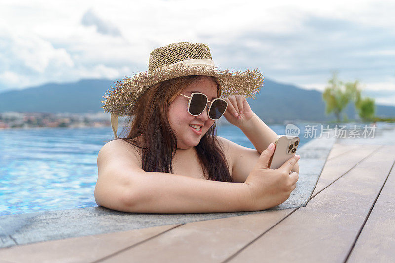 超重的年轻女子橙色swimsuitÂ在游泳池里放松地拿着智能手机。肥胖女人度假旅行用手使用手机