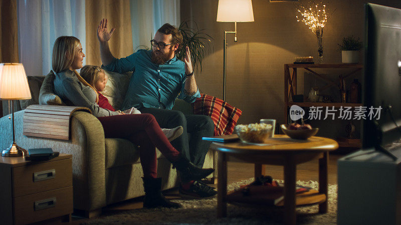父亲，母亲和小女孩看电视的可能性不大。他们坐在舒适的客厅里的沙发上吃着爆米花。这是晚上。