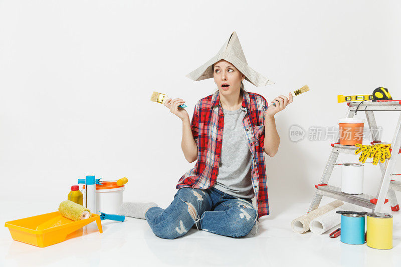 有趣的女人戴着报纸帽坐在地板上用刷子刷，仪器为装修公寓房间隔离在白色的背景上。墙纸，胶水配件，油漆工具。修理家里的概念。