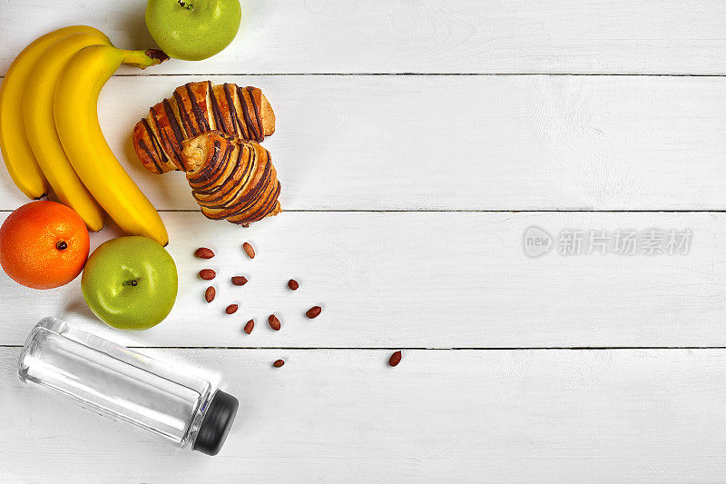 水果早餐，木桌上有空余空间。羊角面包，香蕉，苹果，坚果和一瓶水。俯视图