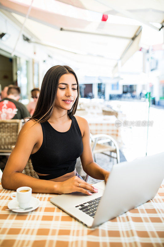 夏日里，年轻的女学生坐在咖啡馆里，呼吸着新鲜空气，一边在笔记本电脑上敲击键盘，一边喝咖啡。漂亮的拉丁女人在早餐时通过上网本连接无线网络