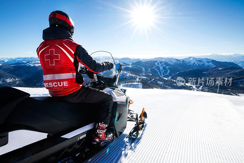 在一个阳光明媚的日子里，搜救人员坐在他的机动雪橇上，看着美丽的山腰景色