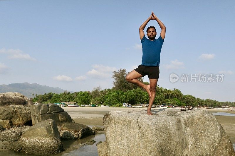 印度人在岩石上练习树姿势(Vrksasana)瑜伽姿势，水的边缘，帕洛伦海滩，果阿，印度