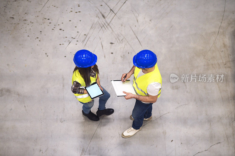 两名土建工程师站在灰色混凝土表面，用平板和图纸高角度观察施工现场讨论项目。