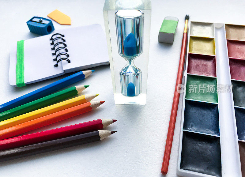 沙漏的特写和文具学校和创意，绘画和工艺品(水彩颜料，彩色铅笔，橡皮，卷笔刀)与白色纹理粗糙的水彩纸