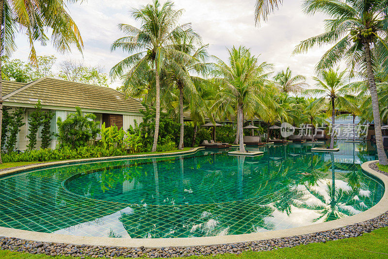 在海边游泳池度假村和酒店，游泳池的风景环绕椰子树和小屋。泳池边度假平房的游客在Hushin泰国