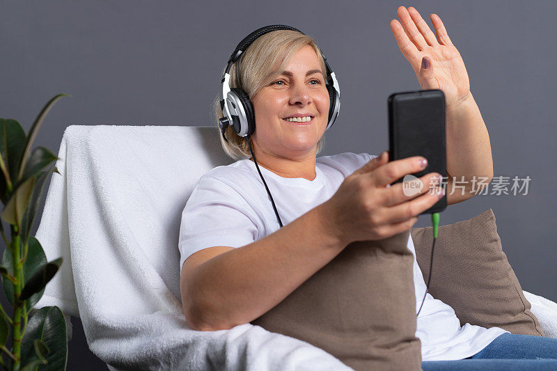 一位年长的女士坐在舒适的椅子上，戴着耳机打视频电话。高级在线概念和大流行概念。